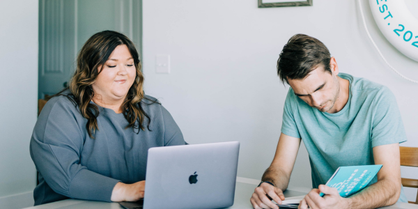 Two people sitting next to each other, one in front of a laptop and one holding a book